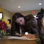 Highlands Latin School student writes her name in book on table with flowers, grapes, vines, and columns around