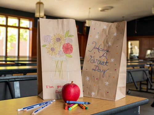 2 brown paper bags with different colorful drawings and phrases on them display on a table with an apple, crayons, and colored pencils in front of them