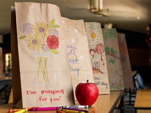 Brown paper bag with a mason jar and flowers drawn on sits on a table with crayons, an apple, and other brown paper bags surrounding it